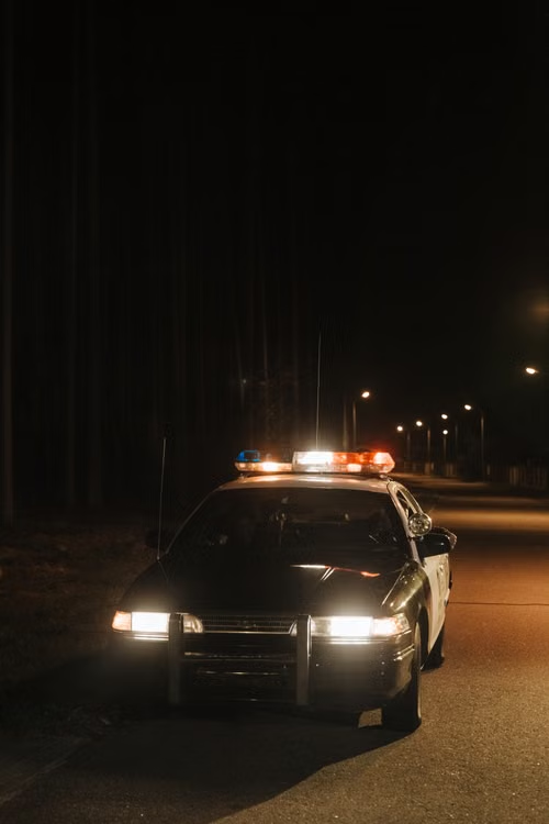 A solitary car parked on a quiet, dark street at night with emergency lights flashing.